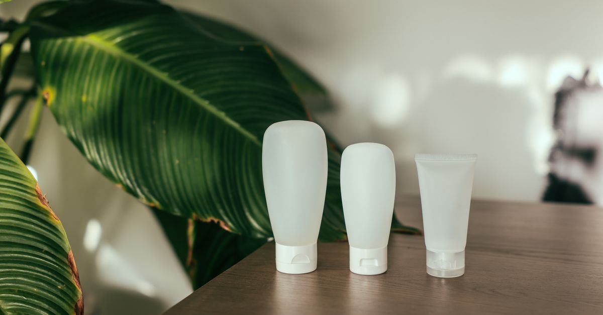 Three plain lotion bottles on a wooden table beside green leaves