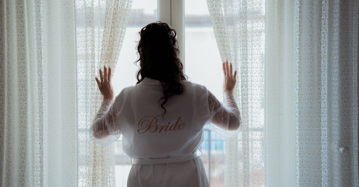 Bride in a robe looking out a bright window on her wedding morning